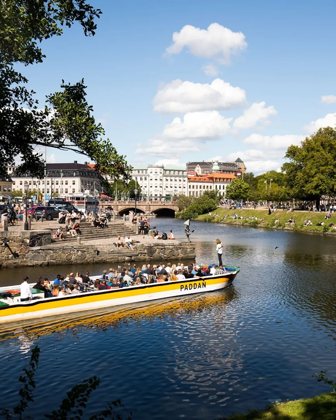 A flat-bottomed Paddan sightseeing boat packed with tourists passing under a very low bridge on Gothenburg's canals, showing historic buildings and waterfront life.